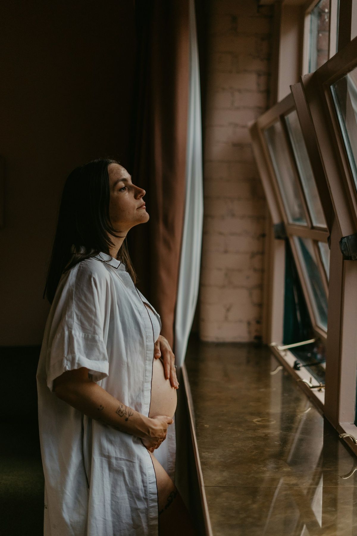 Gravid kvinne i hvit skjorte ved vinduet hjemme